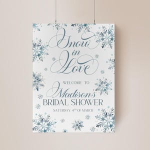 May include: A white poster with the words "Snow in Love" in elegant script, welcoming guests to Madison's Bridal Shower. The design features watercolor snowflakes in shades of blue and gray, with the date "Saturday, 4th of March" at the bottom.