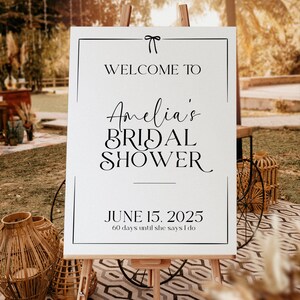 May include: A white welcome sign with black text and a black border. The text reads "Welcome to Amelia's Bridal Shower June 15, 2025 60 days until she says I do".