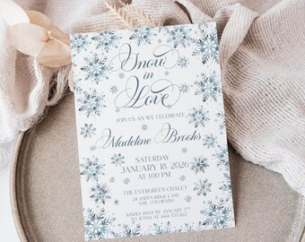 May include: A wedding invitation with the words "Snow in Love" and snowflake illustrations. The invitation includes the names "Madeline Brooks", the date, time, and location of the event. The invitation is on a neutral background.