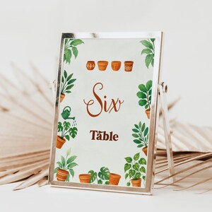 May include: A silver framed table sign with a botanical theme. The sign features the word "Six Table" in a decorative font, surrounded by watercolor illustrations of potted plants and greenery. The background is a soft, off-white color.