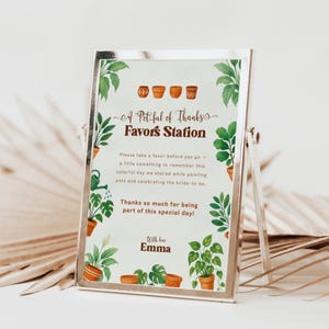 Puede incluir: Un letrero con marco plateado con el texto "A Pot-ful of Thanks Favors Station". El letrero tiene ilustraciones de acuarela de plantas en macetas y el texto "With love Emma". El letrero está sobre una superficie blanca.