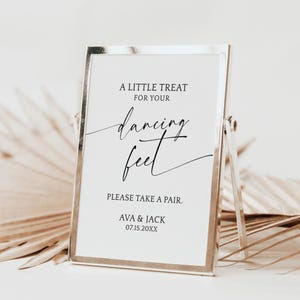 May include: A framed sign with the text "A LITTLE TREAT FOR YOUR dancing feet PLEASE TAKE A PAIR. AVA & JACK 07.15.20XX". The sign has a silver frame and is propped up by a stand.