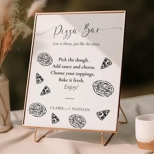 以下が含まれることがあります： 金色の枠と黒いスタンドが付いた白い看板。看板には「Pizza Bar」と書かれており、かわいいピザのイラストが描かれている。テキストには「Love is cheesy, just like this pizza. Pick the dough. Add sauce and cheese. Choose your toppings. Bake it fresh. Enjoy! CLAIRE & NATHAN」と書かれている