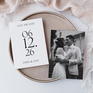 Könnte beinhalten: Ein Hochzeitskarten-Set mit einer "Save the Date"-Karte mit dem Datum "06.12.26" und den Namen "Emma & Jack". Eine Schwarz-Weiß-Fotokarte zeigt ein küssendes Paar. Die Karten liegen auf einem runden, neutralfarbenen Tablett.