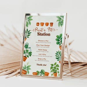 May include: A silver framed sign with the text "Paint a Pot Station" and instructions for a painting activity. The sign is decorated with illustrations of potted plants and a watering can. The sign is supported by a silver stand.
