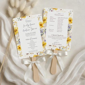 Peut inclure: Deux éventails de mariage avec des motifs floraux. Les éventails ont une poignée en bois et sont attachés avec un ruban blanc. Le texte sur les éventails comprend les noms d'Emily Grace et Andrew James, la date et l'ordre de la cérémonie.