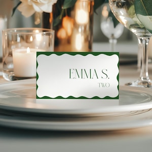 May include: A white place card with a scalloped green border, displaying the name "EMMA S." and the number "TWO" in a dark green font. The card is set on a white plate, with a lit candle and a glass of wine in the background.