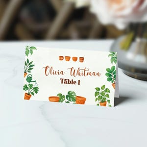 May include: A folded place card with a watercolor design featuring potted plants and greenery. The card displays the name "Olivia Whitman" and "Table 1" in brown script. Decorative terracotta pots are also featured.