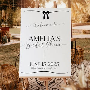 Może przedstawiać: Biały znak z czarnym tekstem i czarną kokardką u góry. Tekst brzmi "Witamy na Bridal Shower Amelii | 15 czerwca 2025 60 dni do ślubu".