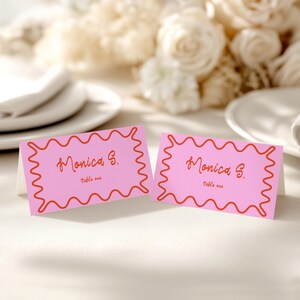 May include: Two pink place cards with a wavy red border and the name "Monica S." printed in a cursive font. The words "table one" are printed below the name. The cards are on a white surface with flowers in the background.
