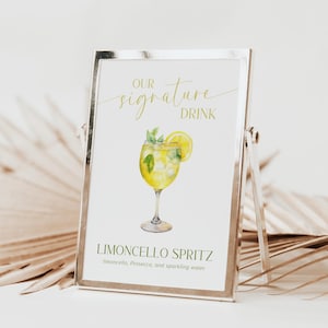 May include: A silver framed sign featuring the words "OUR signature DRINK" in gold script. It displays a watercolour illustration of a Limoncello Spritz cocktail with lemon and mint. The text "LIMONCELLO SPRITZ" is at the bottom.