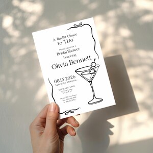 May include: A white invitation card with black text and a martini illustration. The card reads "A Tini Bit Closer to I Do" and invites guests to a bridal shower honouring Olivia Bennett on August 15, 2026.