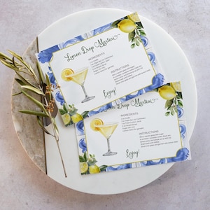 以下が含まれることがあります： レモン ドロップ マティーニのレシピカード2枚。カクテルグラス、レモン、青い花のイラストが描かれています。カードには材料と手順が記載されています。「Enjoy!」の文字が見えます。