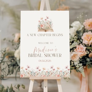 May include: A cream-colored sign on a wooden easel, featuring floral illustrations and text. The sign reads "A New Chapter Begins," "Welcome to Madison's Bridal Shower," and the date 06.14.2026. Floral accents adorn the top and bottom.