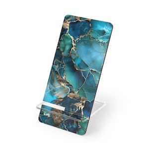 May include: A teal and gold abstract patterned phone stand. The stand has a clear acrylic base and a rectangular top with a marbled design in shades of teal, blue, and gold. The stand is designed to hold a phone or tablet.