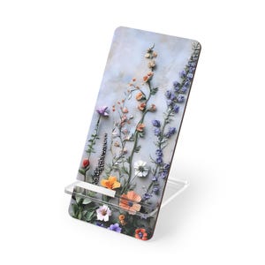 May include: A phone stand made of wood with a floral design. The stand features a variety of flowers in shades of purple, pink, orange, and white on a light blue background.