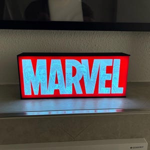 Könnte beinhalten: Ein rechteckiger Leuchtkasten mit dem Wort "MARVEL" in großen, hellblauen Buchstaben. Die Buchstaben sind vor einem leuchtend roten Hintergrund gesetzt. Der Kasten ist schwarz und scheint beleuchtet zu sein.