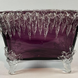 Op de afbeelding: Een rechthoekige, paarse glazen kom met heldere, ijspegelsachtige accenten rond de rand en op de voetjes. De kom heeft een donkerpaars interieur en wordt verhoogd door drie decoratieve voetjes.