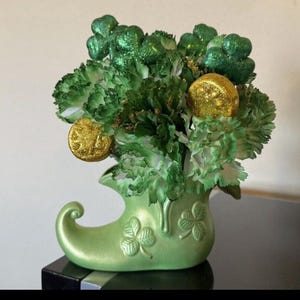 May include: A St. Patrick's Day floral arrangement in a green ceramic leprechaun boot. The arrangement features green carnations, glitter shamrocks, and gold coin accents. The boot has shamrock designs.