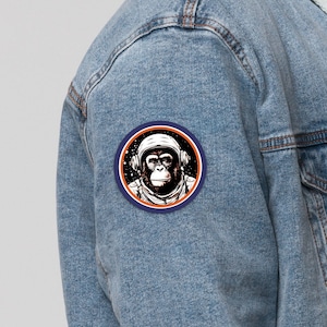 Peut inclure: Une veste en jean bleu clair avec un col en sherpa et un patch représentant un chimpanzé portant un casque d'astronaute. Le patch est sur le côté gauche de la veste.