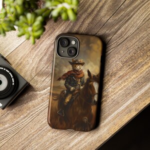 May include: A phone case with a colorful illustration of a cat wearing a cowboy hat and riding a horse. The cat is wearing a brown jacket and a red scarf. The background is a sunset.