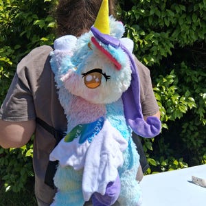 May include: A plush unicorn backpack with a yellow horn, pink and purple mane, and large, expressive eyes. The backpack features a soft, multi-colored design with blue, pink, and white hues, and includes a white, cloud-like detail. The straps are black.