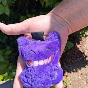 May include: A purple plush toy character with a stitched face, featuring a wide grin with pink teeth and red accents. The toy has a black loop at the top. The character is held in a hand against a backdrop of green leaves.