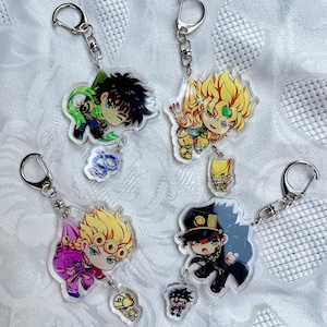 May include: Four anime character keychains with silver key rings. The keychains feature characters from the anime series JoJo's Bizarre Adventure. The characters are Jotaro Kujo, Dio Brando, Giorno Giovanna, and Bruno Bucciarati. Each keychain has a small charm attached to it.