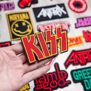 Può includere: Una toppa da stirare ricamata in rosso e giallo con la parola "KISS" in grassetto. La toppa è tenuta in una mano.