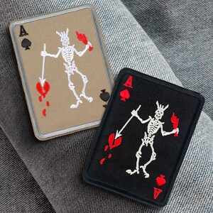 Op de afbeelding: Twee geborduurde patches met de aas van schoppen met een skelet dat een hart en een zwaard vasthoudt. De patches zijn beige en zwart met rode accenten.