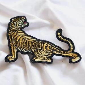 Op de afbeelding: Een geborduurde strijk-op patch met een tijger in goud en zwart. De tijger kijkt naar rechts en heeft een zwart-gouden streepdessin.