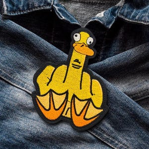 Può includere: Un'anatra gialla che fa il dito medio, ricamata su una giacca di jeans.
