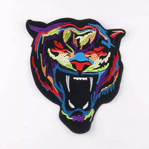 Può includere: Una toppa ricamata colorata con il viso di una tigre con la bocca aperta e denti affilati. La tigre è contornata di nero e presenta una varietà di colori, tra cui rosso, blu, verde, giallo e viola.