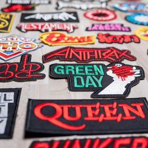 Può includere: Una collezione di toppe ricamate da stirare con vari loghi di band, tra cui Green Day, Queen, Anthrax e The Who. Le toppe sono su uno sfondo di legno chiaro.