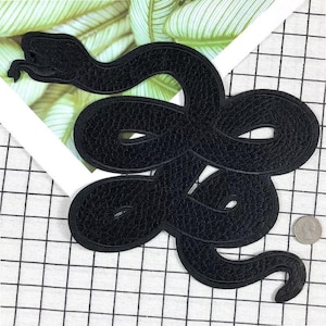 Könnte beinhalten: Schwarzer gestickter Schlangen-Patch. Die Schlange ist zu einem Kreis zusammengerollt, mit dem Kopf nach links gerichtet. Der Patch ist aus einem weichen, strukturierten Stoff.