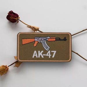 Könnte beinhalten: Ein braunes gesticktes Patch mit einem orangefarbenen AK-47-Gewehr und dem Text "AK-47" in Weiß.