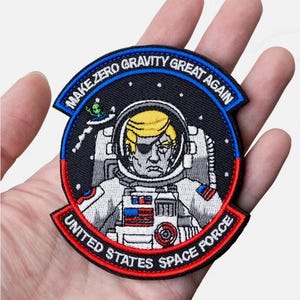 Könnte beinhalten: Gestickter Aufnäher mit schwarzem Hintergrund, der einen Cartoon-Astronauten mit schwarzem Augenklappen zeigt. Der Astronaut trägt einen weißen Raumanzug mit einer amerikanischen Flagge am Arm. Der Text "MAKE ZERO GRAVITY GREAT AGAIN" befindet sich in einem blauen und weißen Bogen über dem Astronauten. Der Text "UNITED STATES SPACE FORCE" befindet sich in einem roten und weißen Bogen unter dem Astronauten.