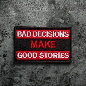 Könnte beinhalten: Roter und schwarzer gestickter Aufnäher mit weißem Text, der "Bad Decisions Make Good Stories" lautet.