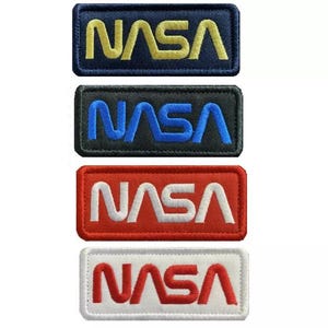 Op de afbeelding: Vier geborduurde patches met het NASA-logo in verschillende kleuren. De patches zijn donkerblauw met gouden letters, zwart met blauwe letters, rood met witte letters en wit met rode letters.