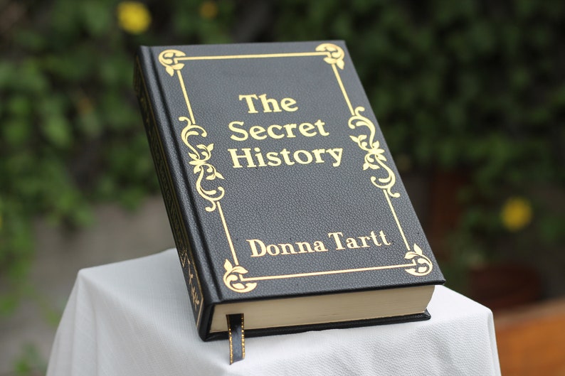 以下が含まれることがあります： 金色の文字で書かれた黒い革装丁の本。タイトルは「The Secret History」で、作者はDonna Tarttです。