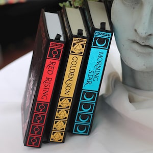 May include: Three hardcover books with black covers and colorful spines. The titles are "Red Rising", "Golden Son", and "Morning Star".
