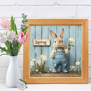 May include: A framed painting of a brown rabbit wearing blue overalls and holding a white flower. The rabbit is standing in front of a blue wooden fence with a sign that says "Spring".