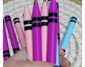 Puede incluir: Una colección de bolígrafos diseñados como ceras de colores, con un cuerpo de plástico liso en tonos rosa, morado y azul claro. Cada bolígrafo tiene dos rayas negras horizontales cerca de la parte superior y una punta dorada.