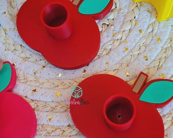 Puede incluir: Dos portavelas con forma de manzana roja con una abertura cilíndrica central y un acento de hoja verde. Cada soporte presenta un pequeño detalle de tallo marrón y un logotipo con el texto "lustraBox" impreso en la superficie de la manzana.