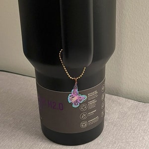 May include: Black stainless steel tumbler with a teal lid and a butterfly charm attached to the handle. The tumbler has a black and white label with the text "H2.0" and information about the product.