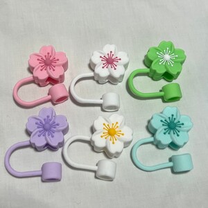 May include: A collection of colorful silicone straw toppers shaped like cherry blossoms. The toppers are in shades of pink, white, green, purple, yellow, and blue. Each topper has a loop and a small cylinder for attaching to a straw.