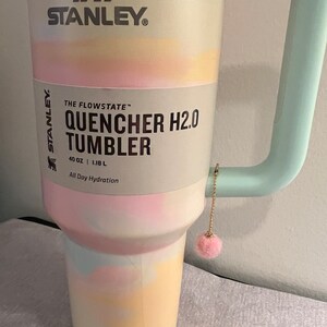 May include: A pastel pink, blue, and yellow Stanley Quencher H2.0 Tumbler with a light blue handle and a pink fuzzy charm. The tumbler is 40 ounces or 1.18 liters and says "All Day Hydration".