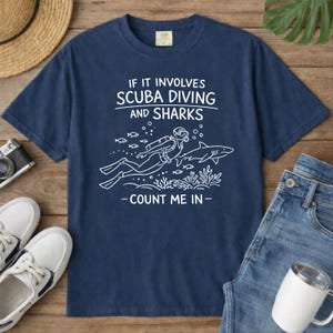May include: Navy blue t-shirt with white text and graphic. The text reads "IF IT INVOLVES SCUBA DIVING AND SHARKS - COUNT ME IN -" with an illustration of a scuba diver and shark. The shirt is paired with jeans, a hat, and shoes.