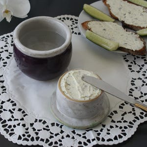 Peut inclure: Un beurrier en céramique avec un couvercle, rempli de beurre aux herbes, et un couteau à beurre. Un petit bol en céramique violet et une assiette de pain beurré et de tranches de concombre sont également présents. Les articles sont sur un napperon blanc.