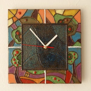 May include: A square ceramic clock with a colourful abstract design. The clock face is dark blue with white hands and red hour hand. The frame is made of colourful ceramic tiles with a mosaic pattern.